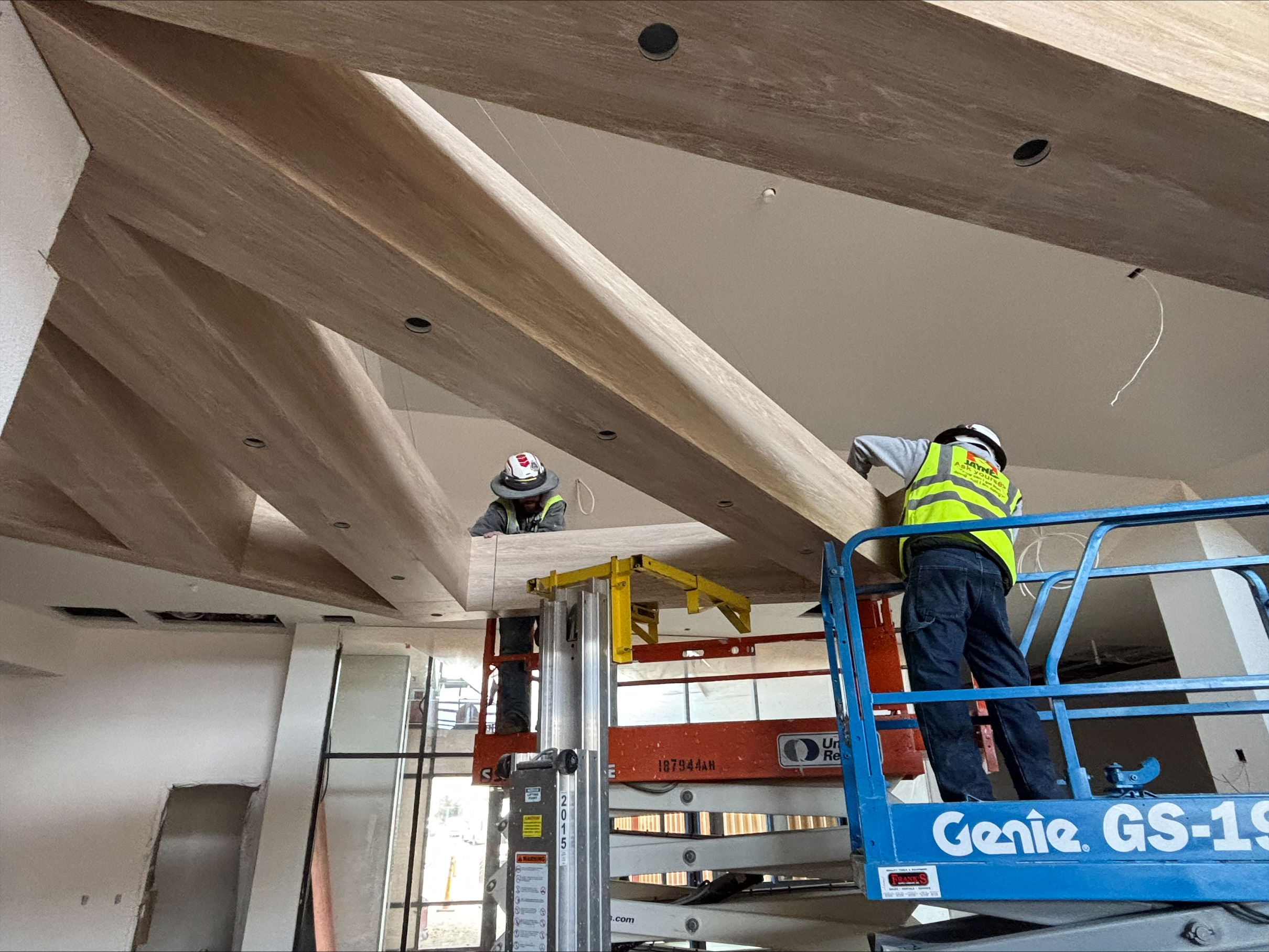 Two carpenters on aerial lifts install large wooden ceiling beams.